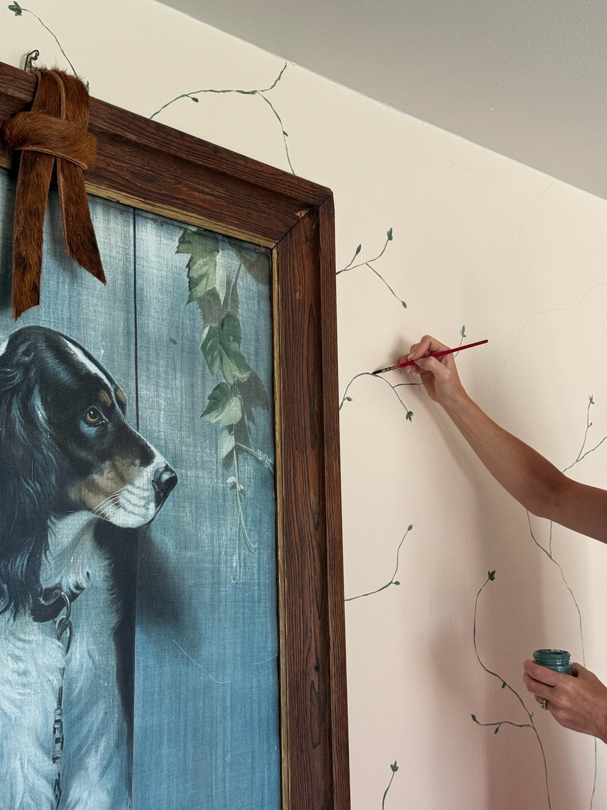 A person's hand and arm are shown painting a vine design onto a light beige wall next to a large framed painting of a black and white dog.